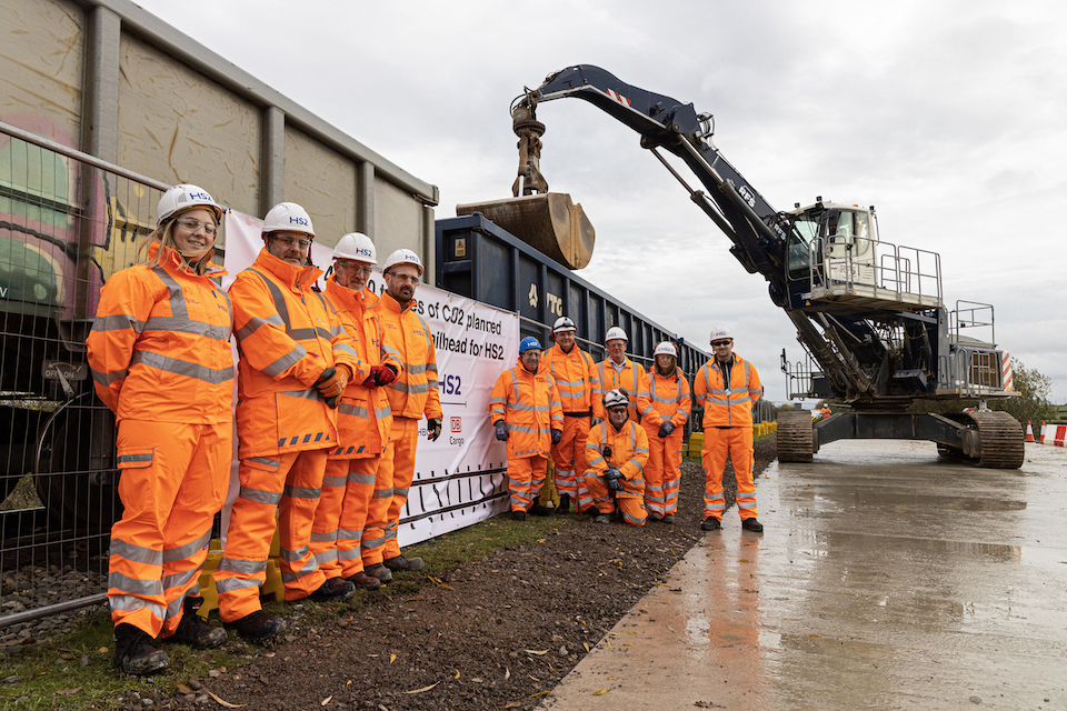 Team of orange suited engineers pose in front of an aggregates train at HS2 Quainton terminal 