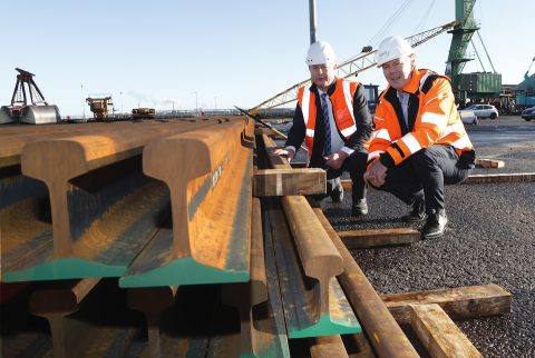 Two men in orange high visibility vests and suits with new rails in a pile in front of them