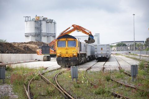 Head on shot of aggregates train being loaded by mechanical shovel with Industrial buildings in the background