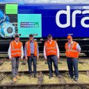 Four gentlemen in orange high visibility vests pose in front of a Drax liveried hopper wagon
