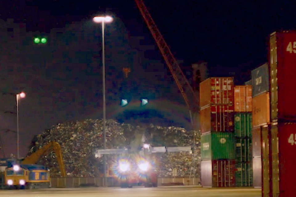 Night time shot of reach stacker with headlights blazing and an intermodal train approaching the terminal
