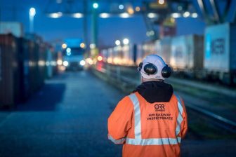 In a high visibility jacket, an inspector from the Office of Rail and Road stands with his back to the camera supervising evening intermodal operations
