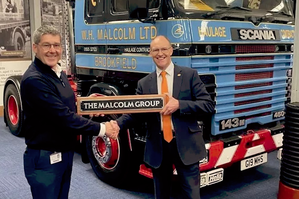 In front of a vintage truck cab, Andrew Malcolm and Tim Shoveller hold a ceremonial plaque which says "The Malcolm Group"