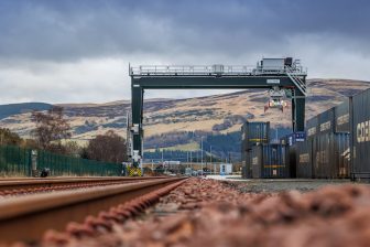 A track level view of the overhead crane at Highland Spring Bottling Plant terminal in Scotland. The red ballast contrasts with the heather on the hills and the grey skies overhead