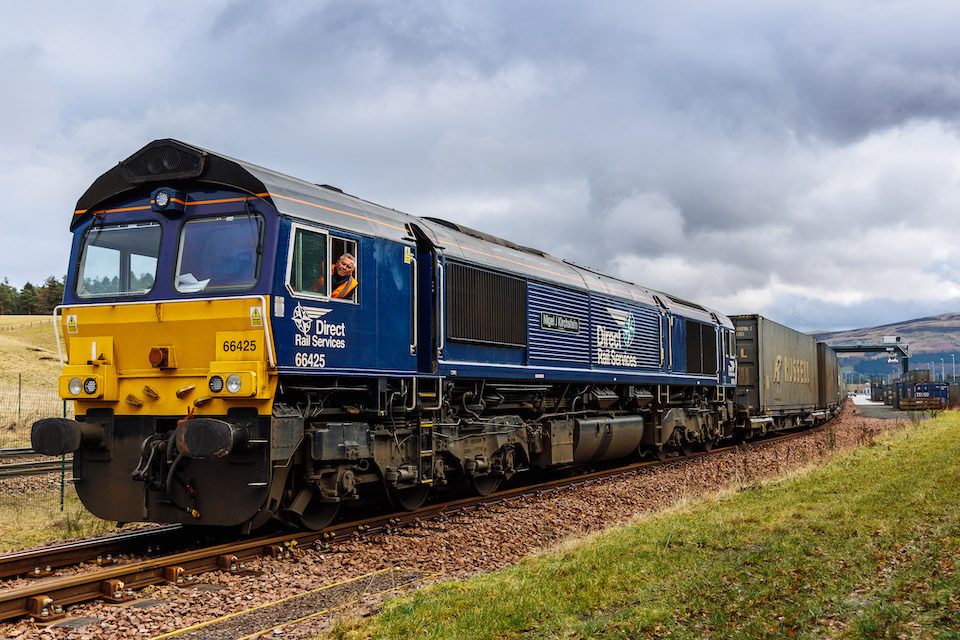 Three quarter view of freight train departing Highland Spring mineral water bottling plant, with the driver looking ou too the window of the class 66 in DRS livery. Cloudy sky