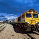 A dusk picture of a Freightliner class 66 diesel at the head of a train of intermodal containers, about to depart a terminal siding under darkening skies