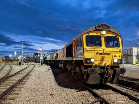 A dusk picture of a Freightliner class 66 diesel at the head of a train of intermodal containers, about to depart a terminal siding under darkening skies