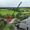 IN a green landscape over a river, a huge red crane lifts an entire bridge structure out over the water